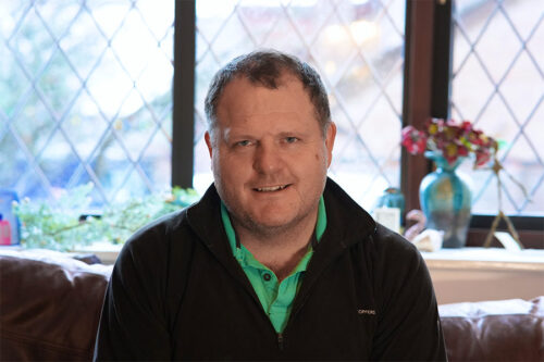 Adam Smith-Connor sits on a couch with a window behind him.